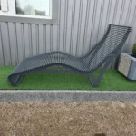 Metāla šezlongs pie ēkas fasādes uz zaļas seguma