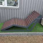 Metāla atpūtas sols ar koka sēdvirsmu sānskatā pie industriālas fasādes