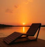 Metāla atpūtas sols ar koka sēdvirsmu saulrieta gaismā pie ūdens